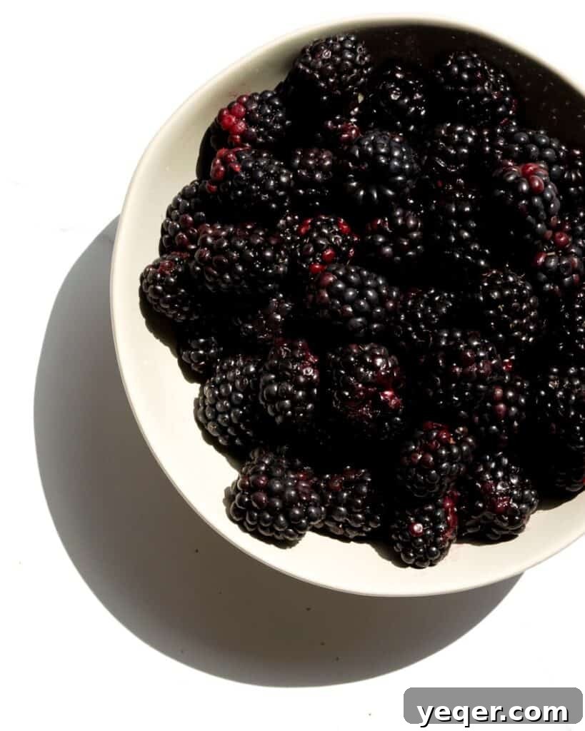 A vibrant bowl overflowing with freshly picked, ripe blackberries sits elegantly on a clean white table, highlighting their rich, dark color and inviting, plump texture. The natural light accentuates the subtle sheen on the berries, making them look irresistible and ready for baking into a delicious summer dessert.