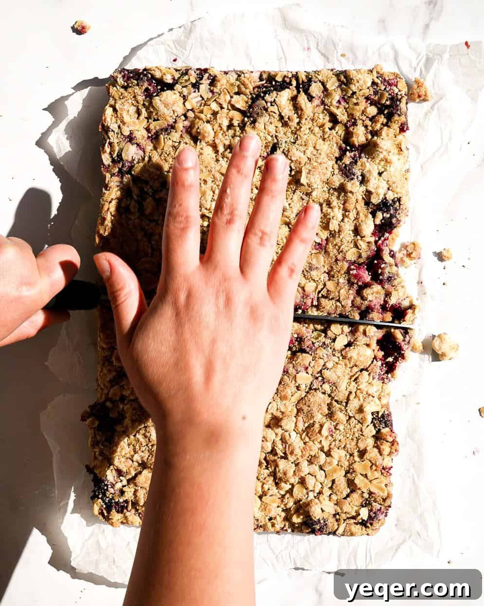 A skilled hand cutting a warm pan of gluten-free blackberry crumb bars into individual, perfect portions using a sharp chef's knife. The steam gently rises, hinting at the fresh, delightful flavors within.