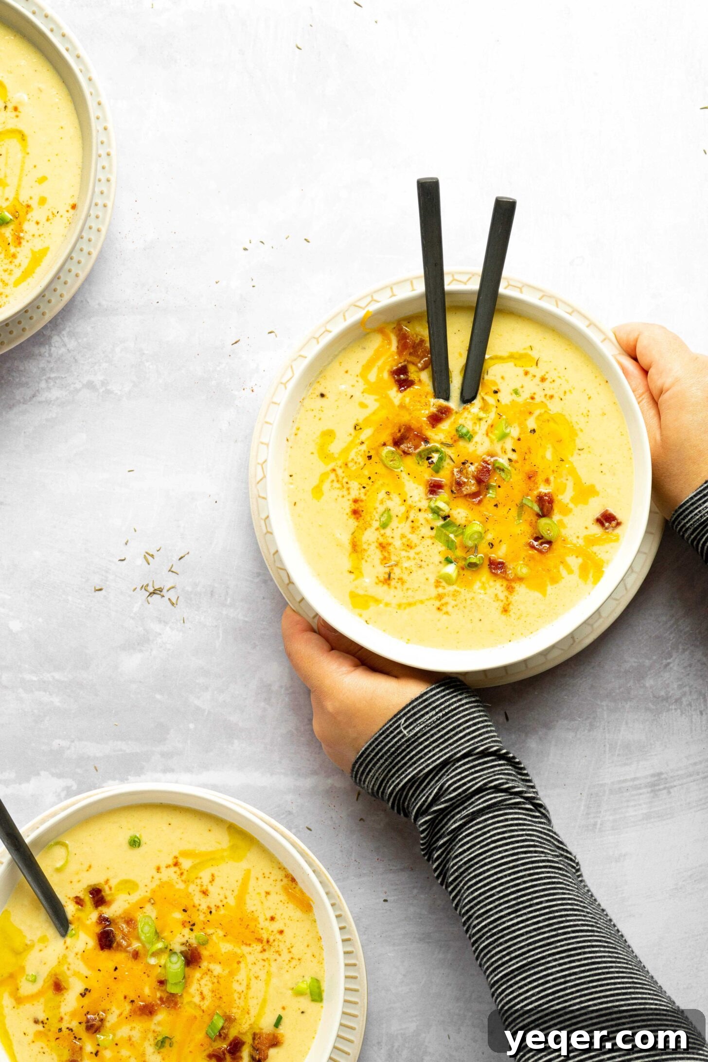 Decadent Loaded Cauliflower Dream 2 Two hands holding a steaming bowl of creamy loaded cauliflower soup, garnished with cheddar cheese, bacon bits, and green onions.