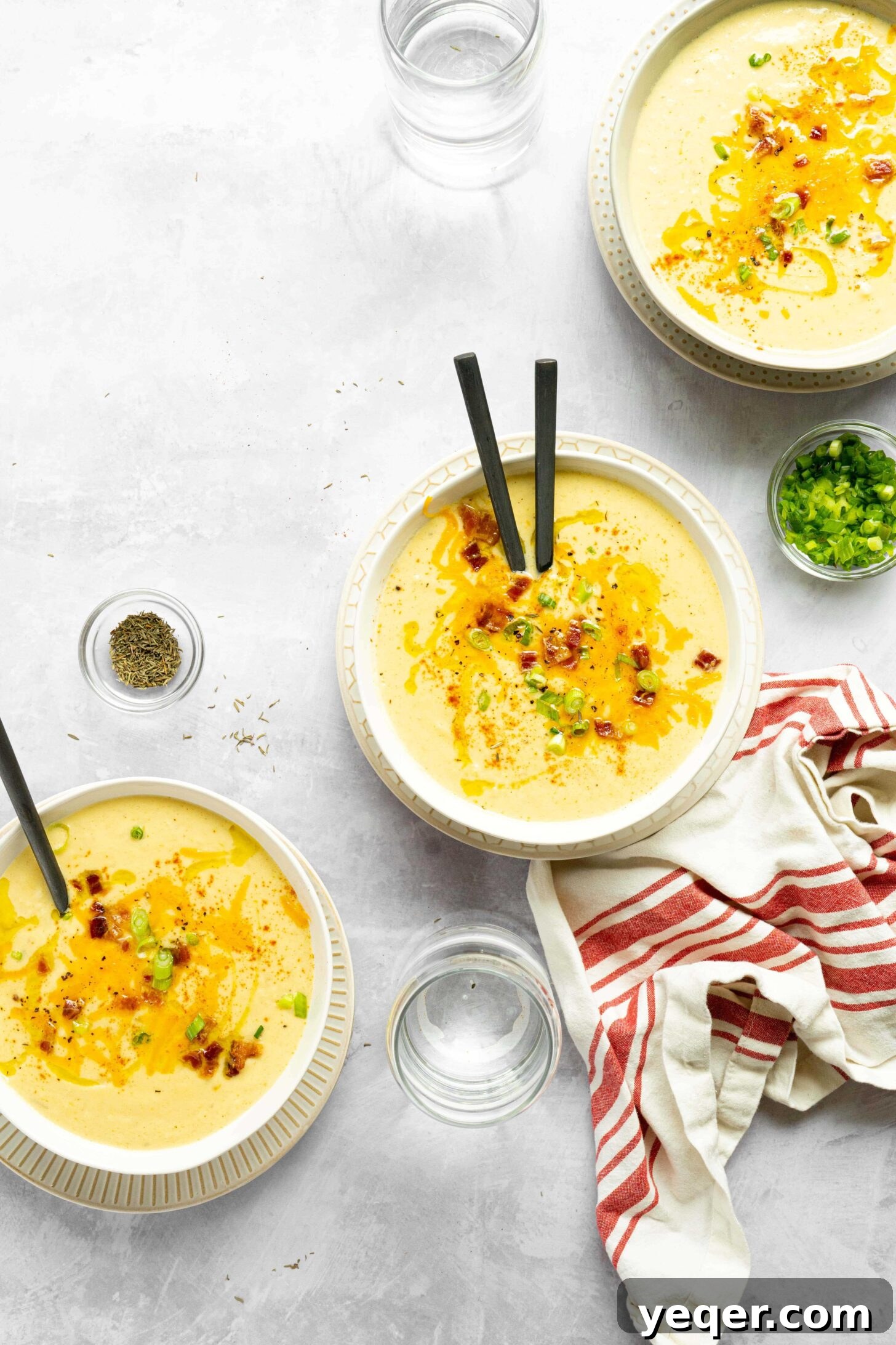 Decadent Loaded Cauliflower Dream 7 Three beautifully presented bowls of loaded cauliflower soup, garnished with bacon, cheese, and green onions, accompanied by water and a napkin.