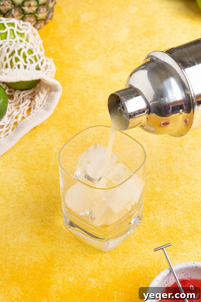 Arctic Tropics 5 A stream of a creamy Pina Colada on the Rocks cocktail being poured from a shaker into a tall glass filled with ice, ready for serving.