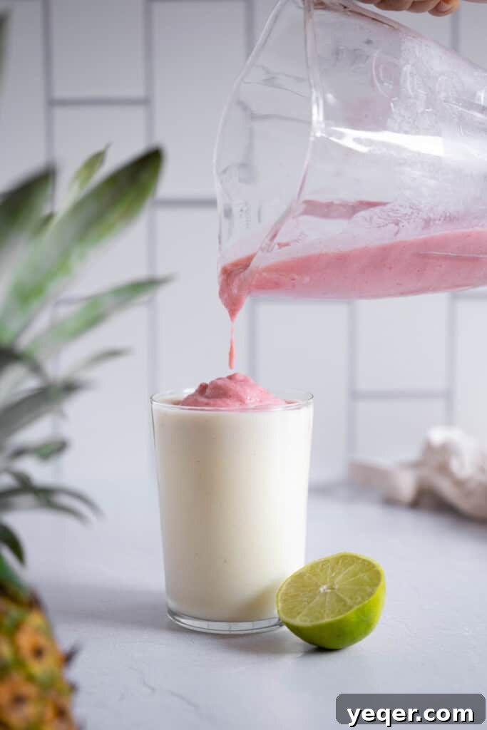 Pink Pina Perfection 7 The vibrant pink strawberry Piña Colada mixture being gracefully poured over the creamy white base layer in a glass, creating a visually captivating and delicious two-toned blended drink.