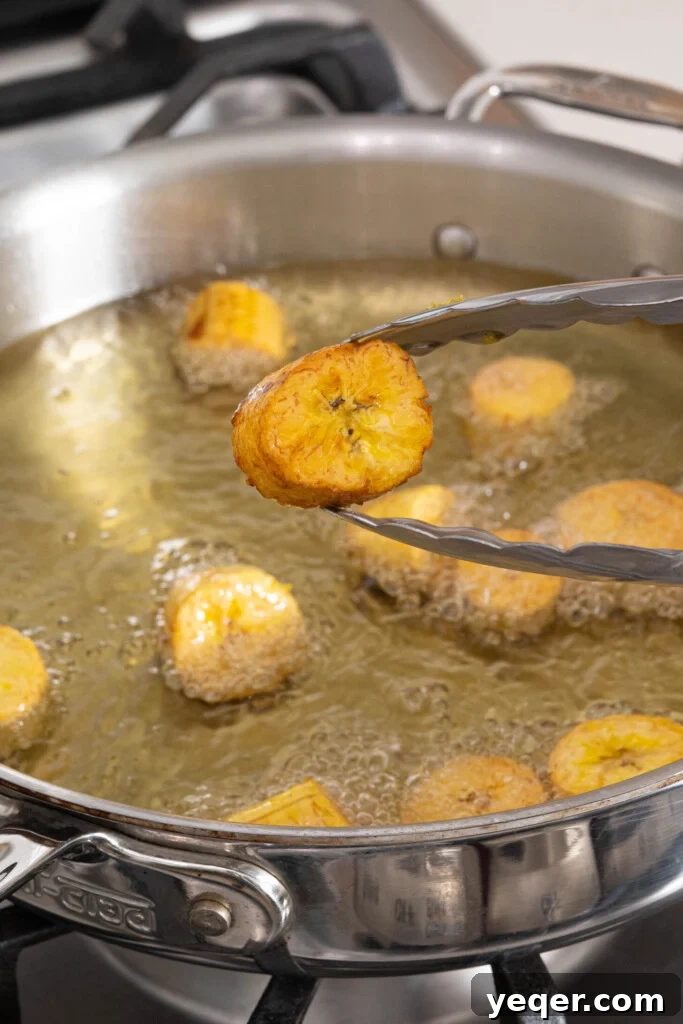 25 Dakikada Geleneksel Porto Riko Mofongosu 6 Tongs carefully holding a freshly fried plantain slice, golden brown and ready for mashing.