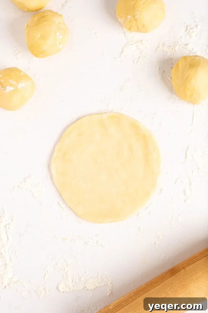 Five-Ingredient Wonder Empanada Dough 11 A close-up shot of hands skillfully rolling out a small ball of empanada dough into a perfect circle with a rolling pin on a floured surface.