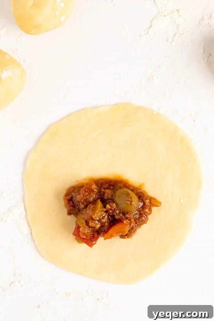 Five-Ingredient Wonder Empanada Dough 12 A hand carefully placing a generous spoonful of picadillo filling onto a round of empanada dough, preparing it for folding and sealing.