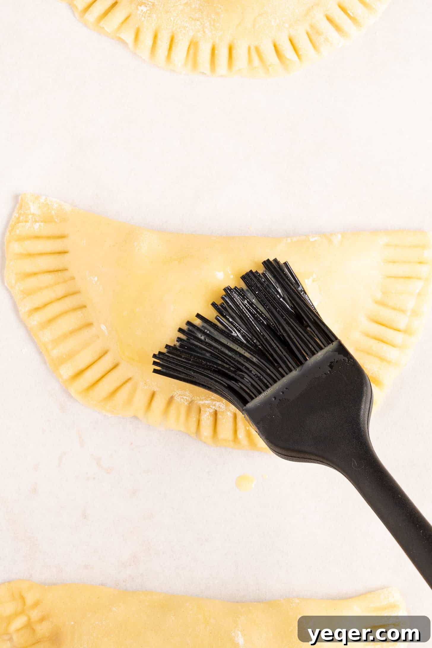 Five-Ingredient Wonder Empanada Dough 18 A pastry brush applies a shiny egg wash to the surface of an uncooked empanada, preparing it for baking and a golden finish.