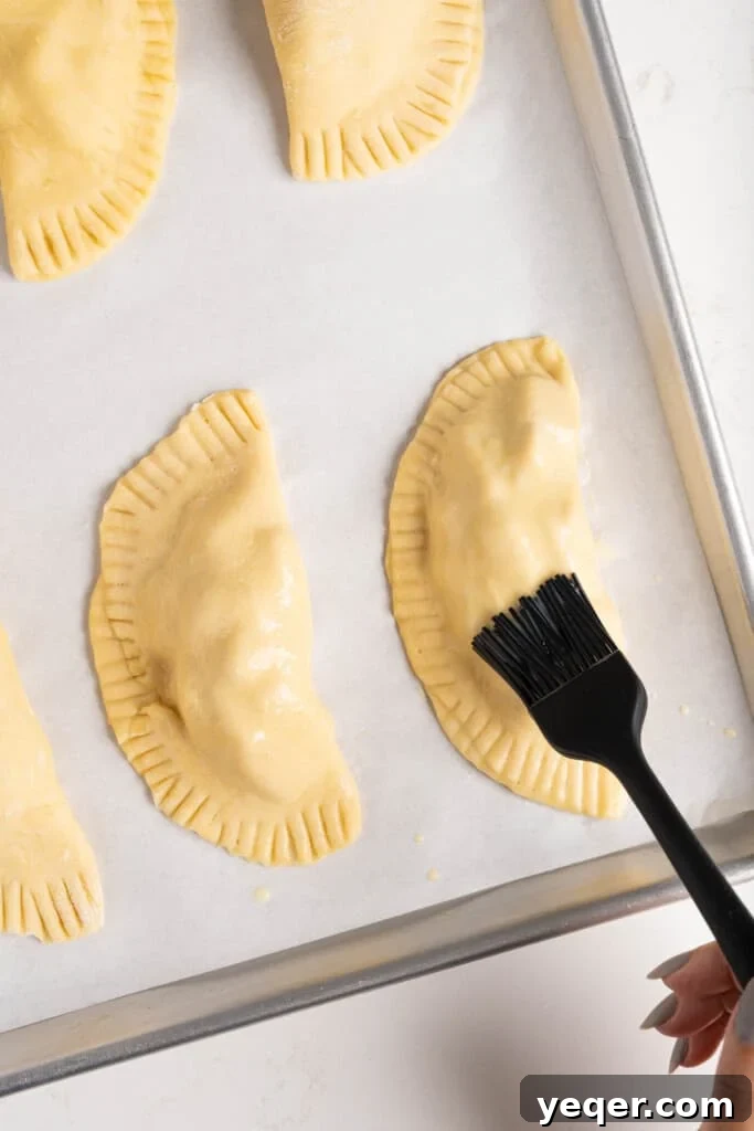 Golden Apple Pastry Pockets 8 Empanadas arranged on a baking sheet, being brushed with an egg wash.