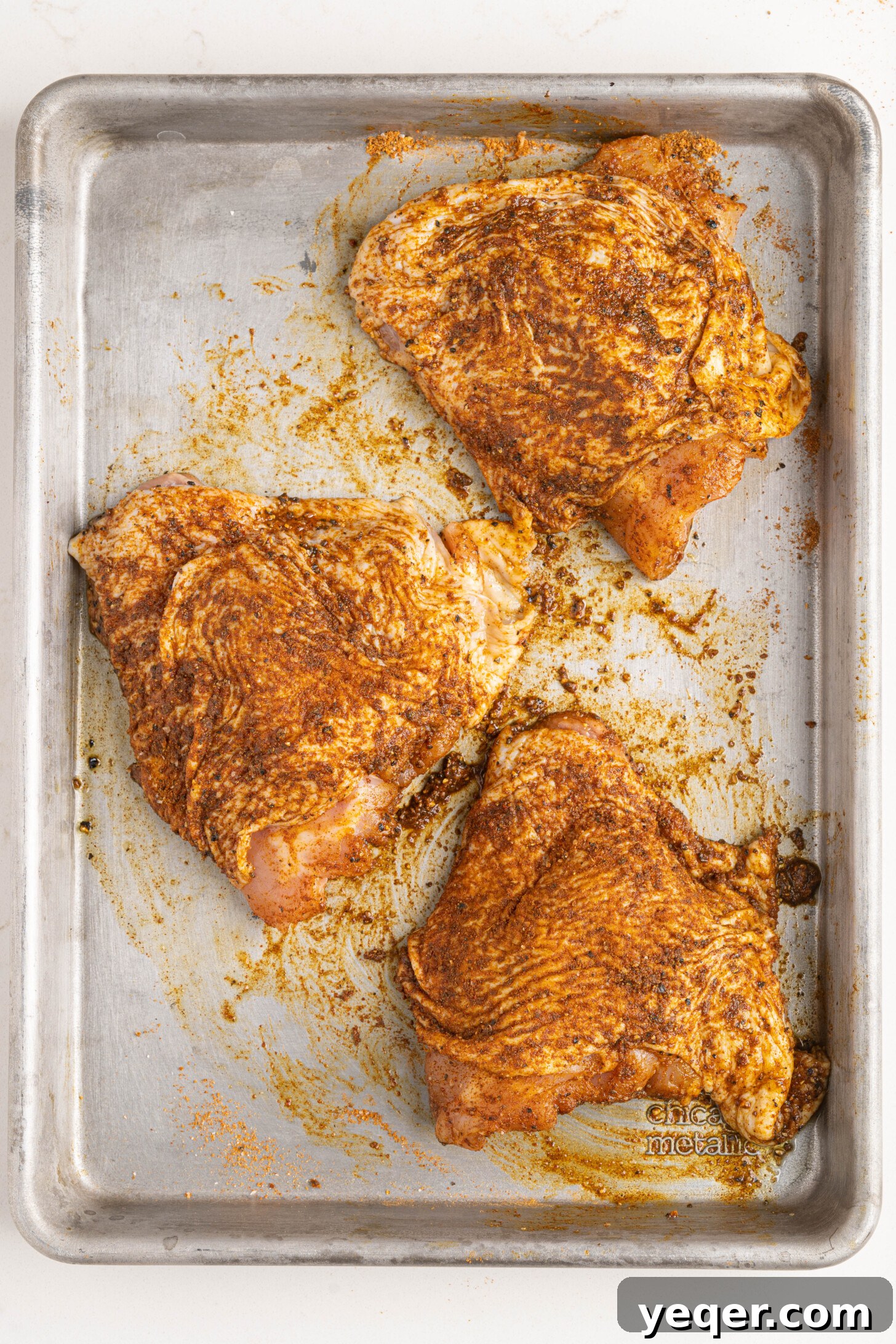 Close-up of chicken thighs generously coated in Ras el Hanout seasoning and olive oil on a baking pan.