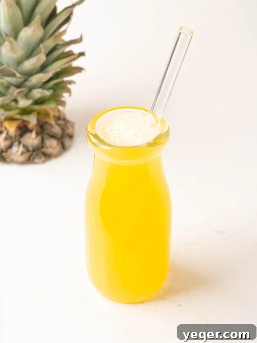 A glass bottle filled with golden homemade pineapple juice, adorned with a straw and a fresh pineapple slice, on a light background.
