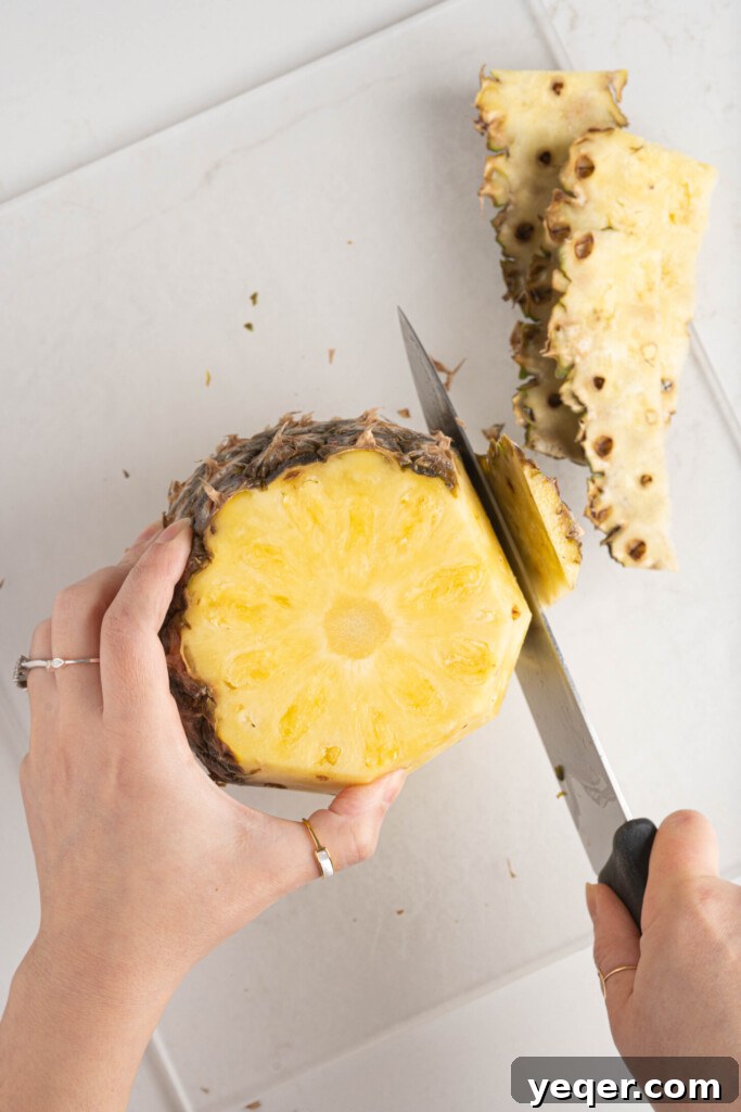 A hand expertly peeling the tough skin off a fresh pineapple on a wooden cutting board with a sharp knife.