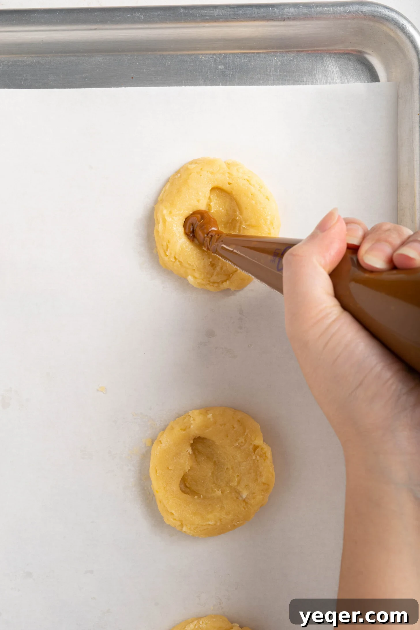 Easy Dulce de Leche Thumbprint Cookies 8 Hands piping creamy dulce de leche into heart-shaped indentations of unbaked sugar cookies on a baking sheet.