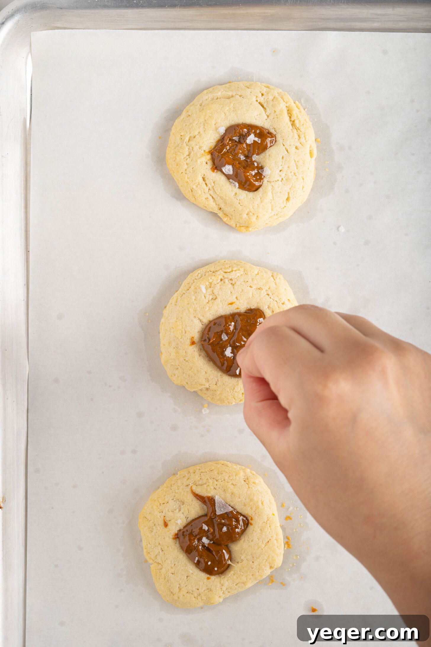 Easy Dulce de Leche Thumbprint Cookies 9 A hand sprinkling flaky sea salt over freshly baked Dulce de Leche cookies, glistening with caramel, on a cooling rack.