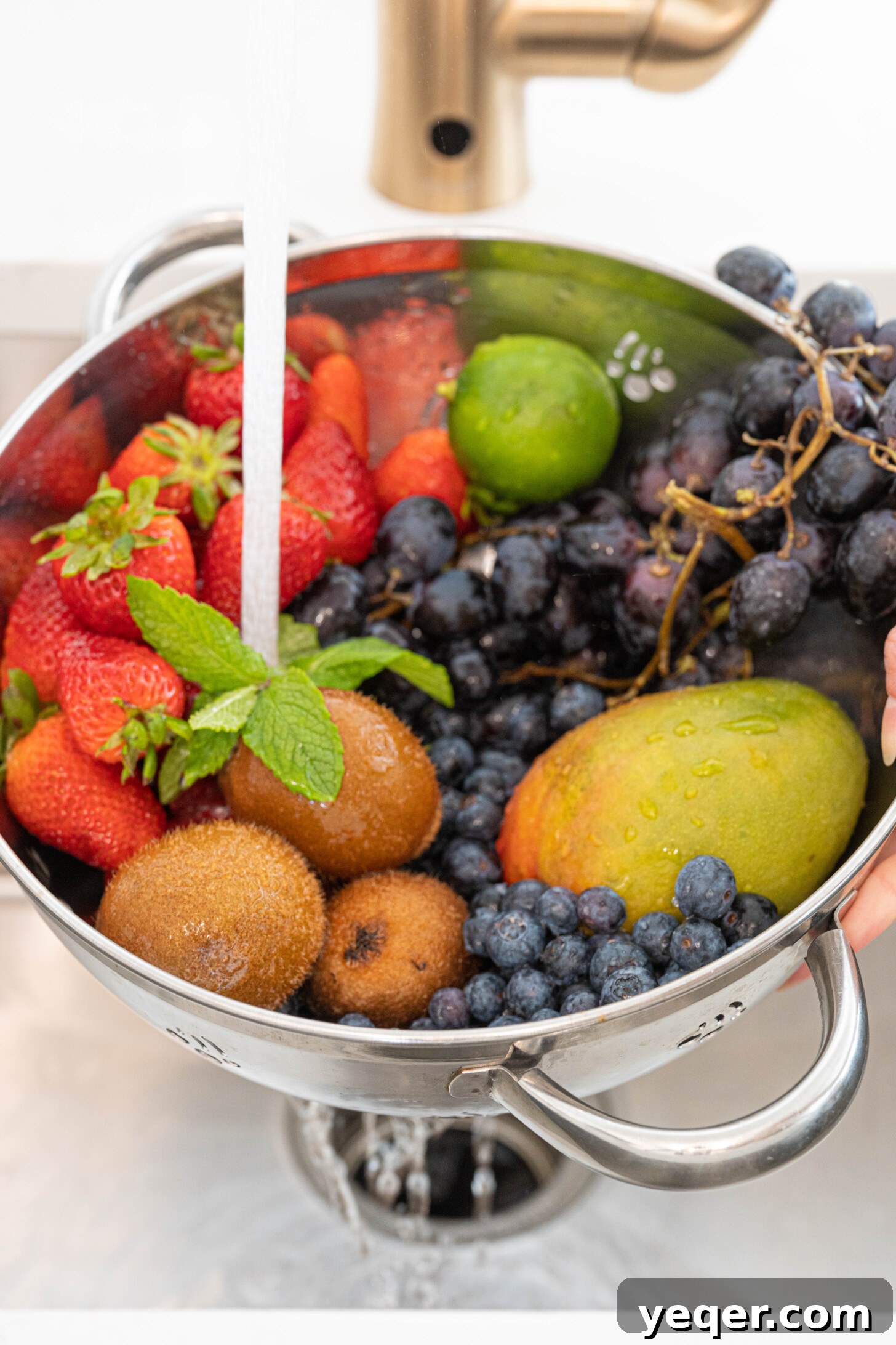 A colander filled with various fruits being rinsed under running water in a kitchen sink, emphasizing the importance of cleanliness.