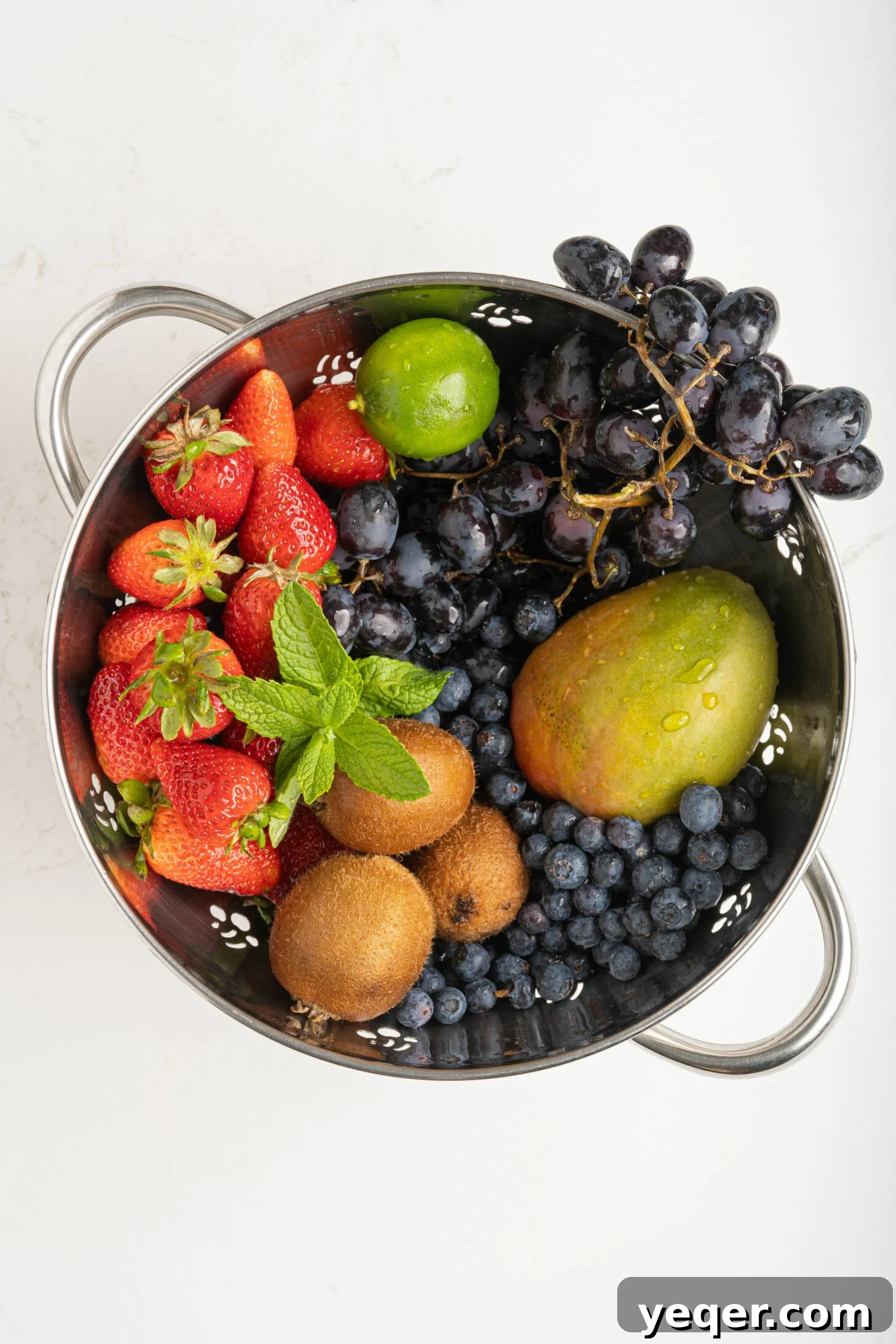 A colander filled with an assortment of fresh fruits including grapes, kiwis, mangoes, strawberries, and blueberries, with fresh mint sprigs nestled among them, ready for preparation.