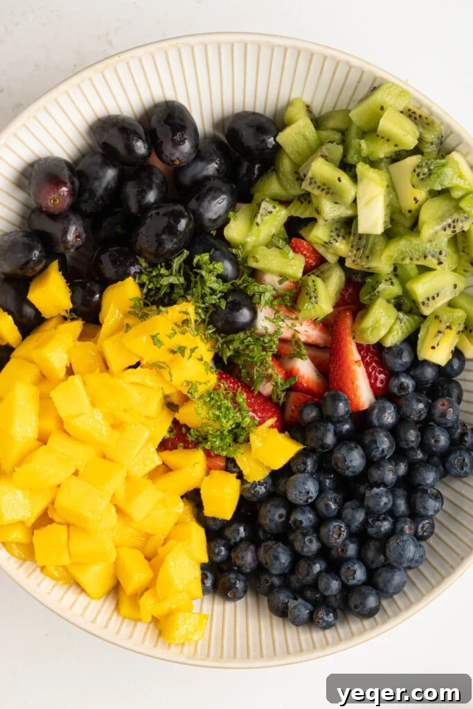 A large bowl containing an assortment of sliced and diced fruits along with lime zest and minced mint, ready to be tossed with lime juice.