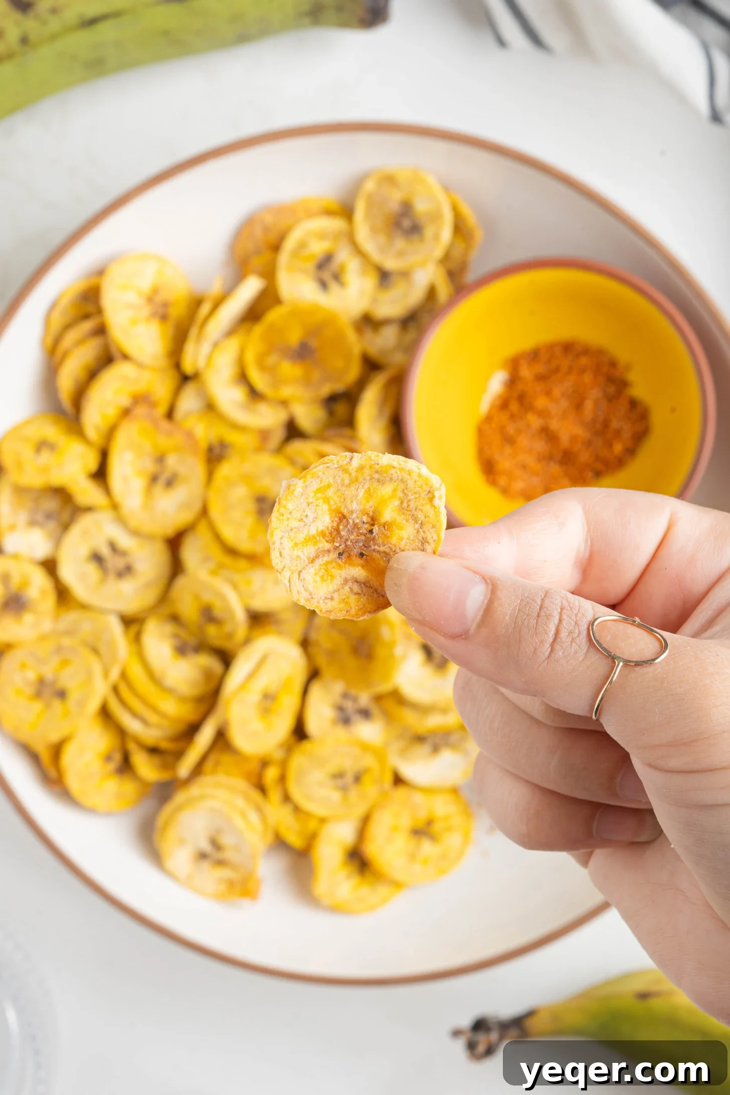 Effortless Oven-Baked Plantain Crisps 12 A hand holds up a single, perfectly golden and crispy baked plantain chip, showcasing its thinness and appetizing texture.