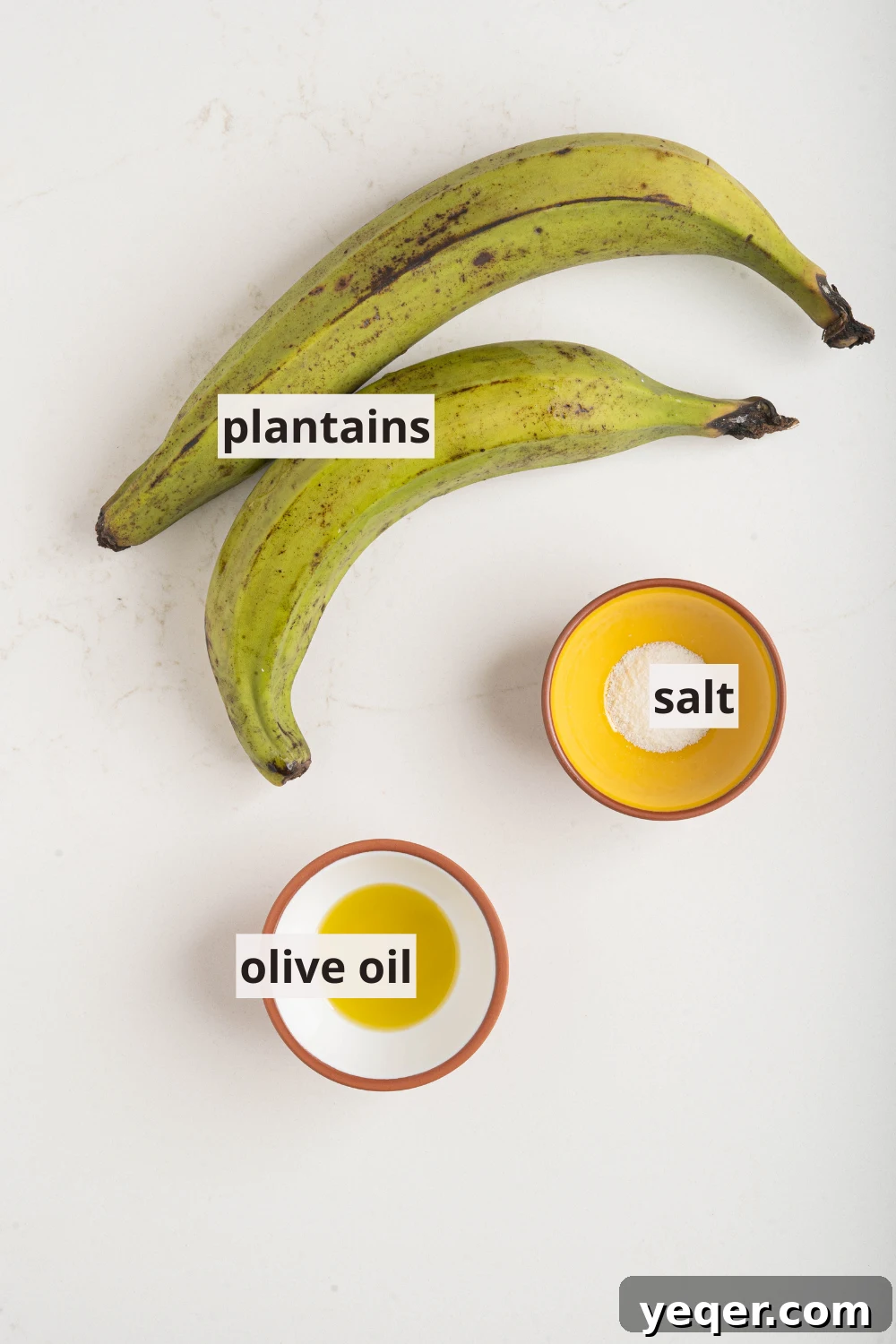 Effortless Oven-Baked Plantain Crisps 3 Raw green plantains, a bowl of coarse salt, and a bottle of olive oil laid out on a clean white kitchen counter, symbolizing the simple ingredients for homemade plantain chips.