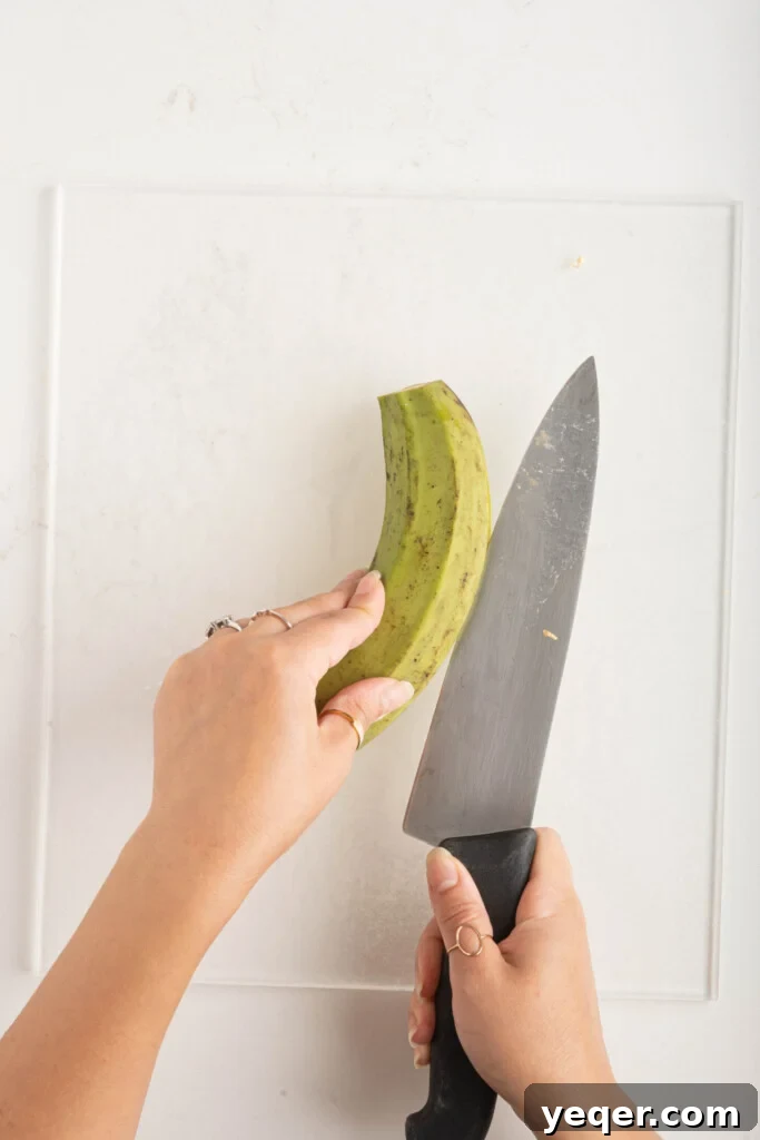 Effortless Oven-Baked Plantain Crisps 4 A knife carefully slicing the peel of a green plantain on a wooden cutting board.