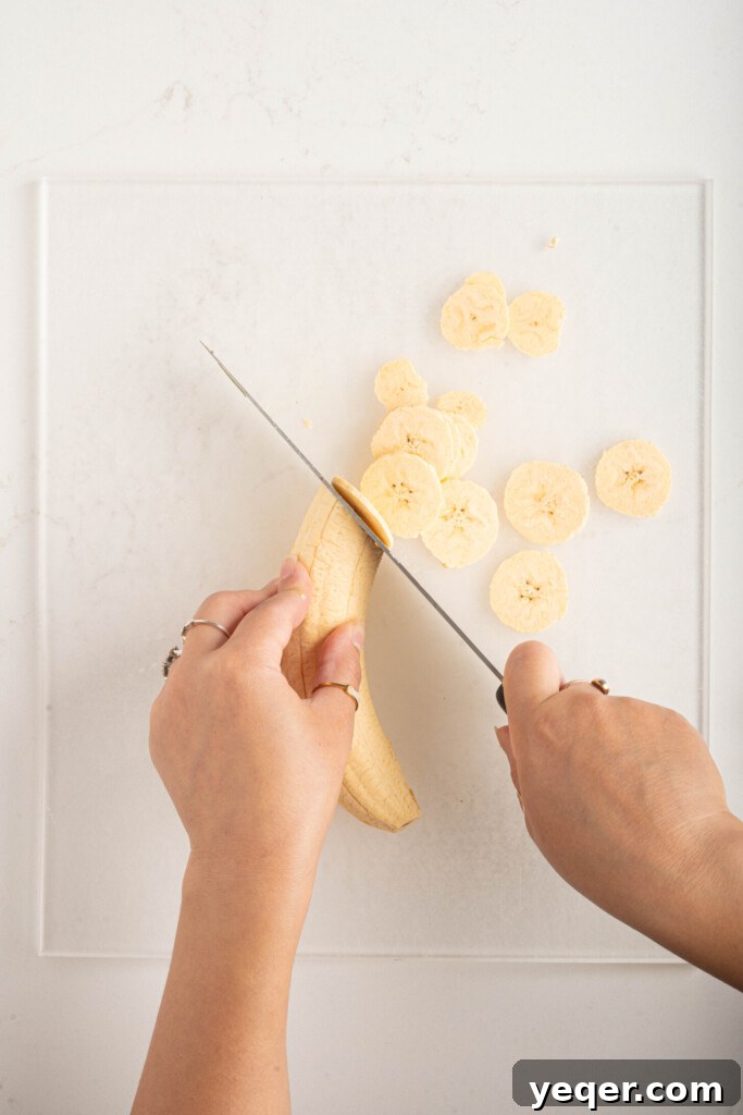 Effortless Oven-Baked Plantain Crisps 6 Hands carefully slicing a peeled green plantain into thin, uniform rounds on a cutting board using a sharp knife.