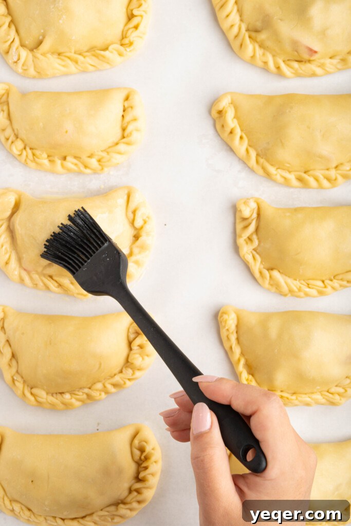 Homestyle Shredded Chicken Empanadas 9 A pastry brush applying a glistening egg wash over the surface of unbaked empanadas on a baking sheet.
