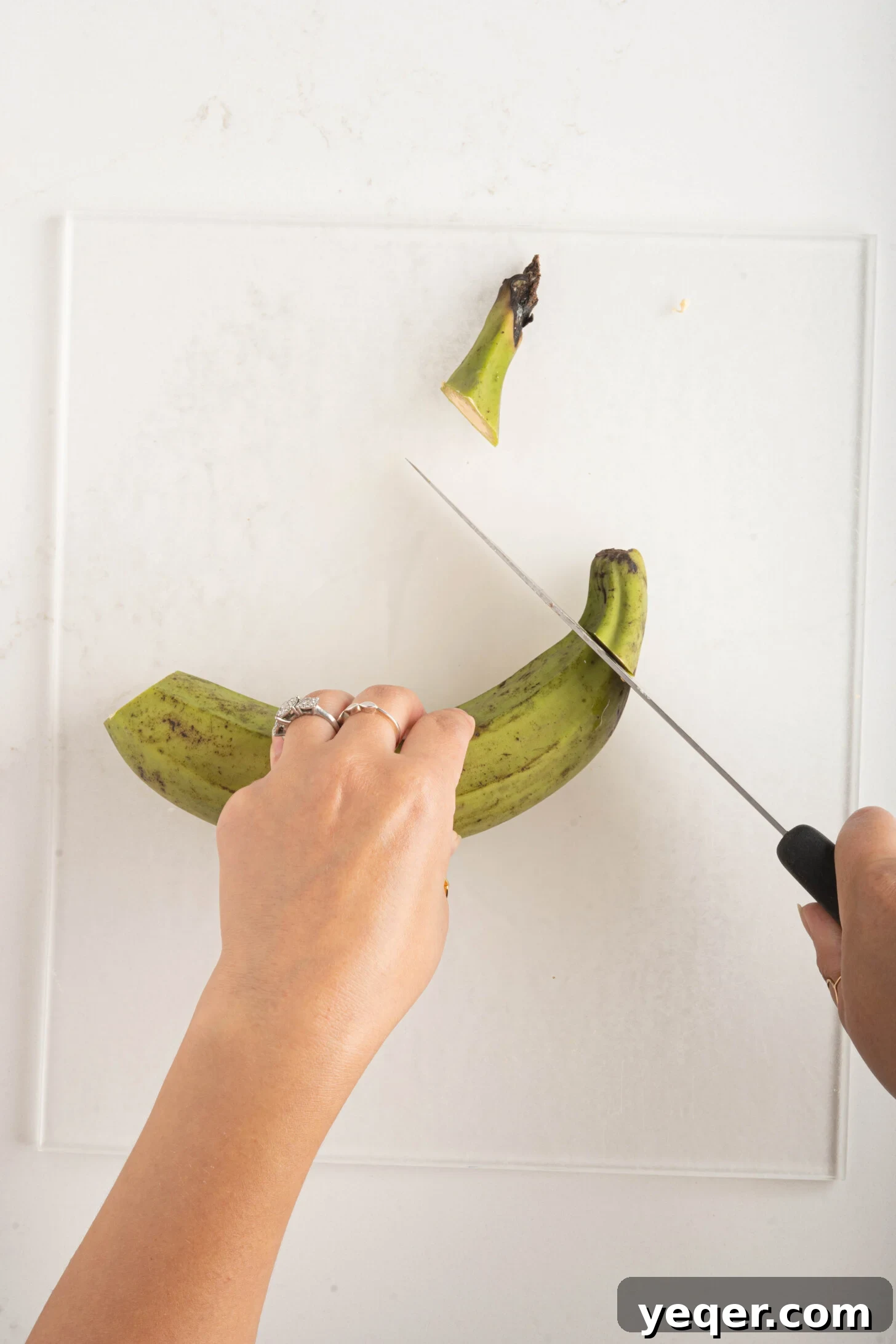 Master the Art of Peeling Any Plantain 7 A pair of hands expertly cutting a green plantain with a knife on a wooden cutting board.