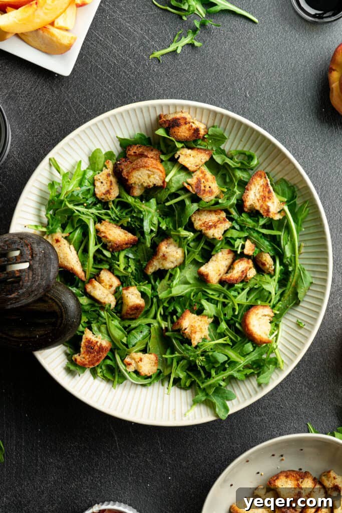 Decadent Peach Prosciutto Burrata Salad with Arugula and Crispy Croutons 5 Arugula with croutons and dressing in a large white salad bowl, showing the initial layering.