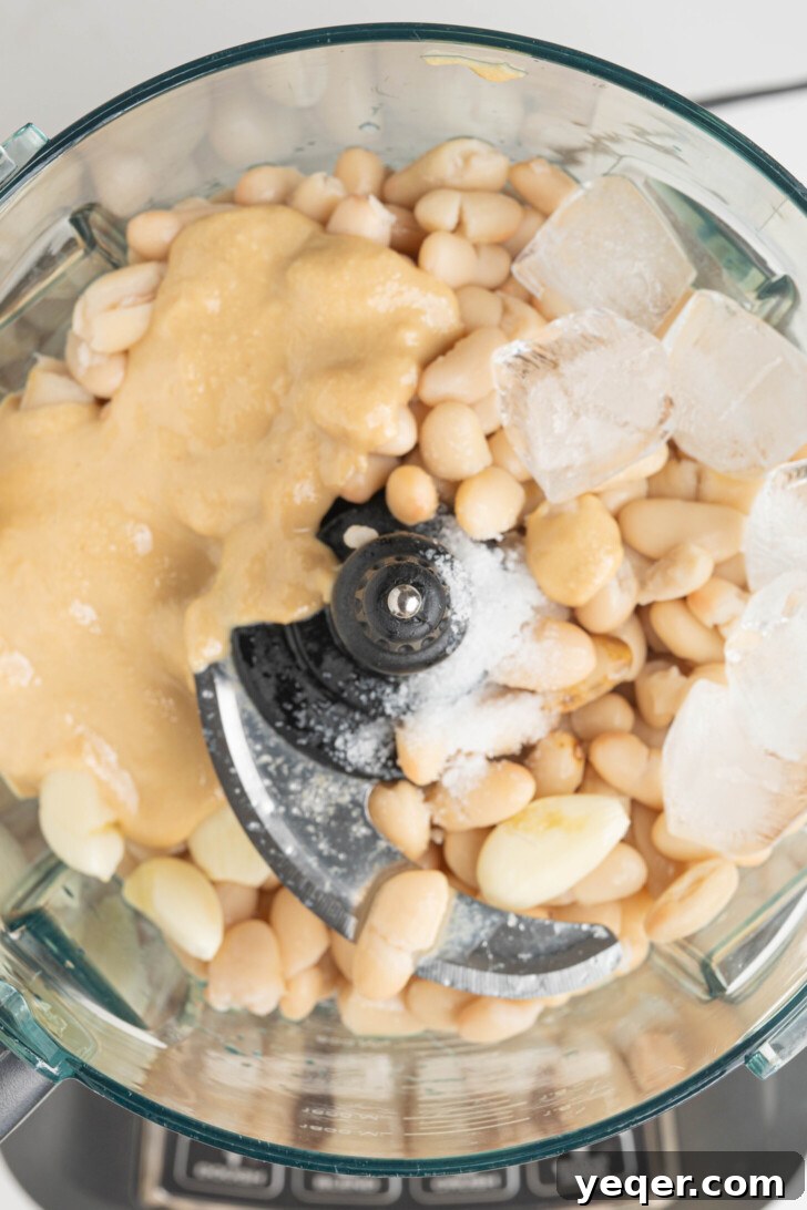 White beans, tahini, ice, and more in a food processor, ready for blending.