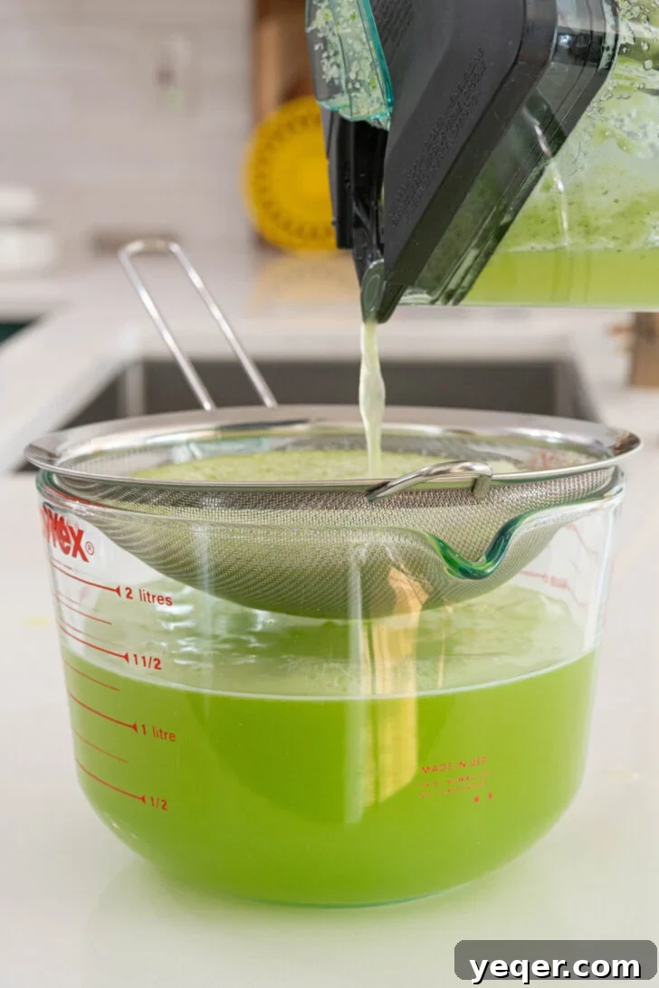 Cool Cucumber Lime Refresher 6 Agua de pepino being strained through a fine-mesh sieve into a large glass pitcher, separating solids from the liquid.