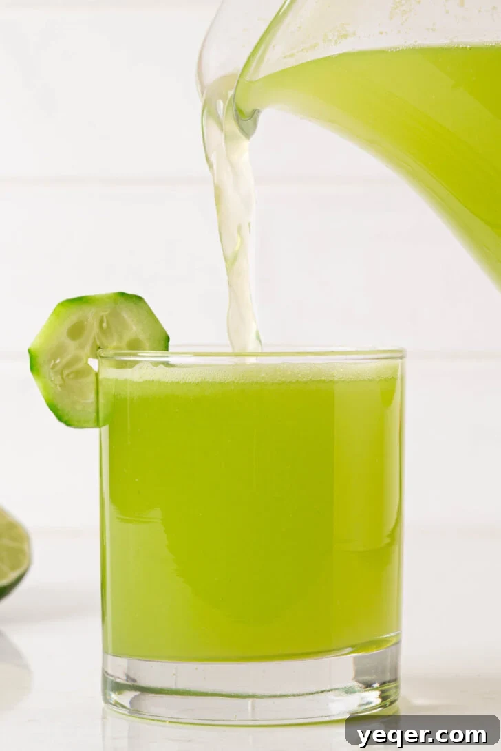 Cool Cucumber Lime Refresher 7 A hand pouring a glass of vibrant green agua de pepino con limon over ice, garnished with fresh cucumber and lime slices.