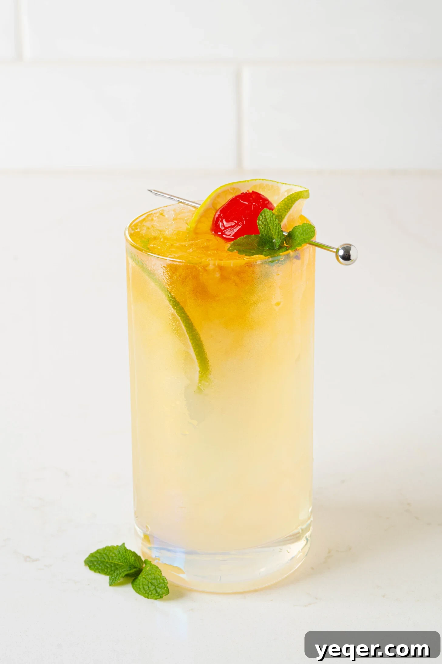 Authentic Mai Tai Cocktail 2 A Mai Tai cocktail, garnished with mint and a lime wedge, sitting on a white counter with a tropical background slightly blurred.
