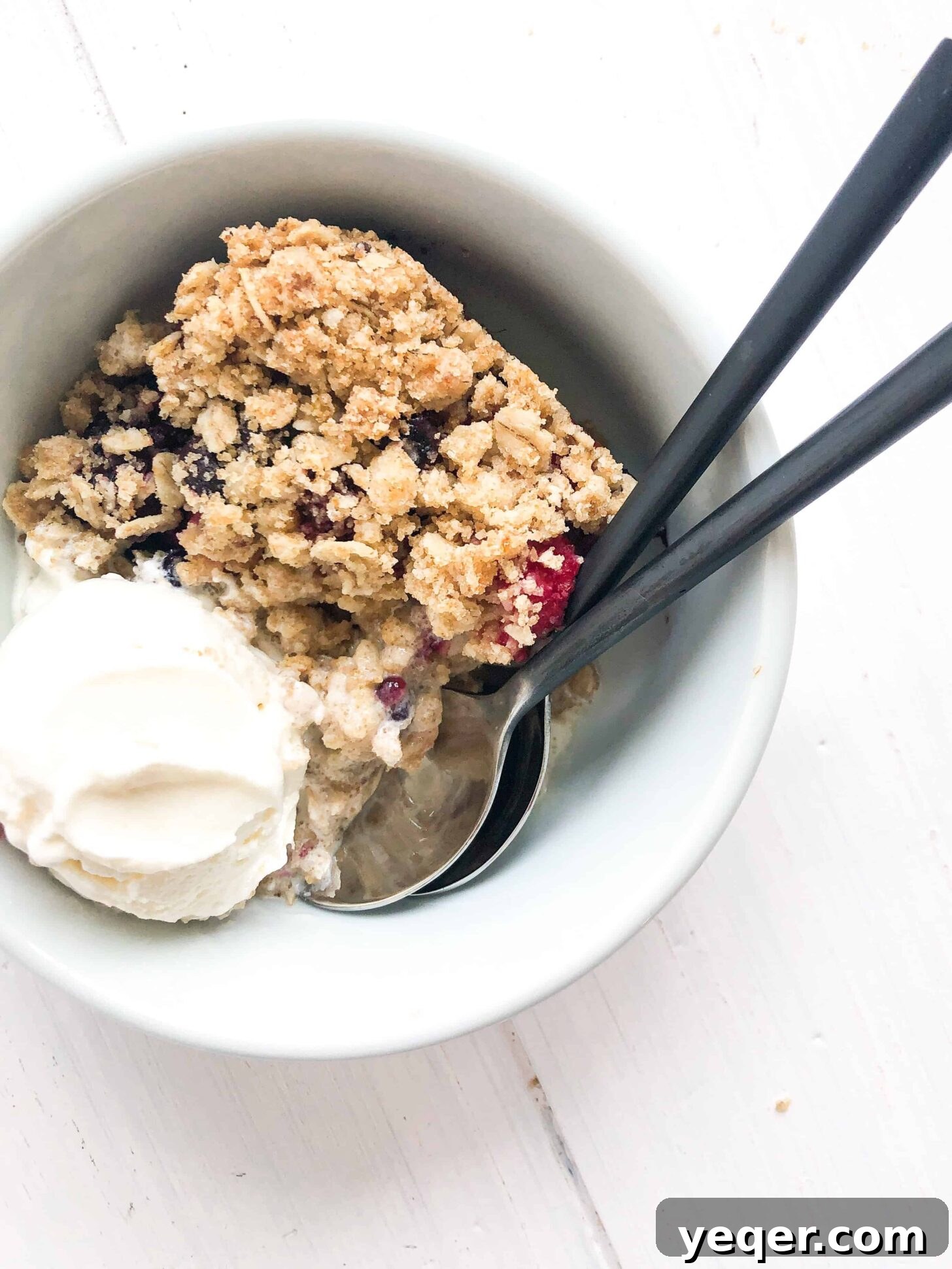 Heavenly Gluten Free Berry Crumble 5 A warm gluten-free berry crisp served in a bowl with two spoons, topped with melting vanilla ice cream, indicating it's ready to be shared and enjoyed.