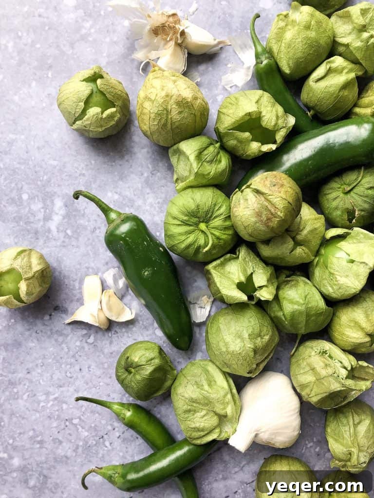 Smoky Tomatillo Salsa 3 A vibrant assortment of fresh ingredients laid out, including green tomatillos, fiery serrano peppers, potent jalapeños, and unpeeled garlic cloves, ready for roasting.